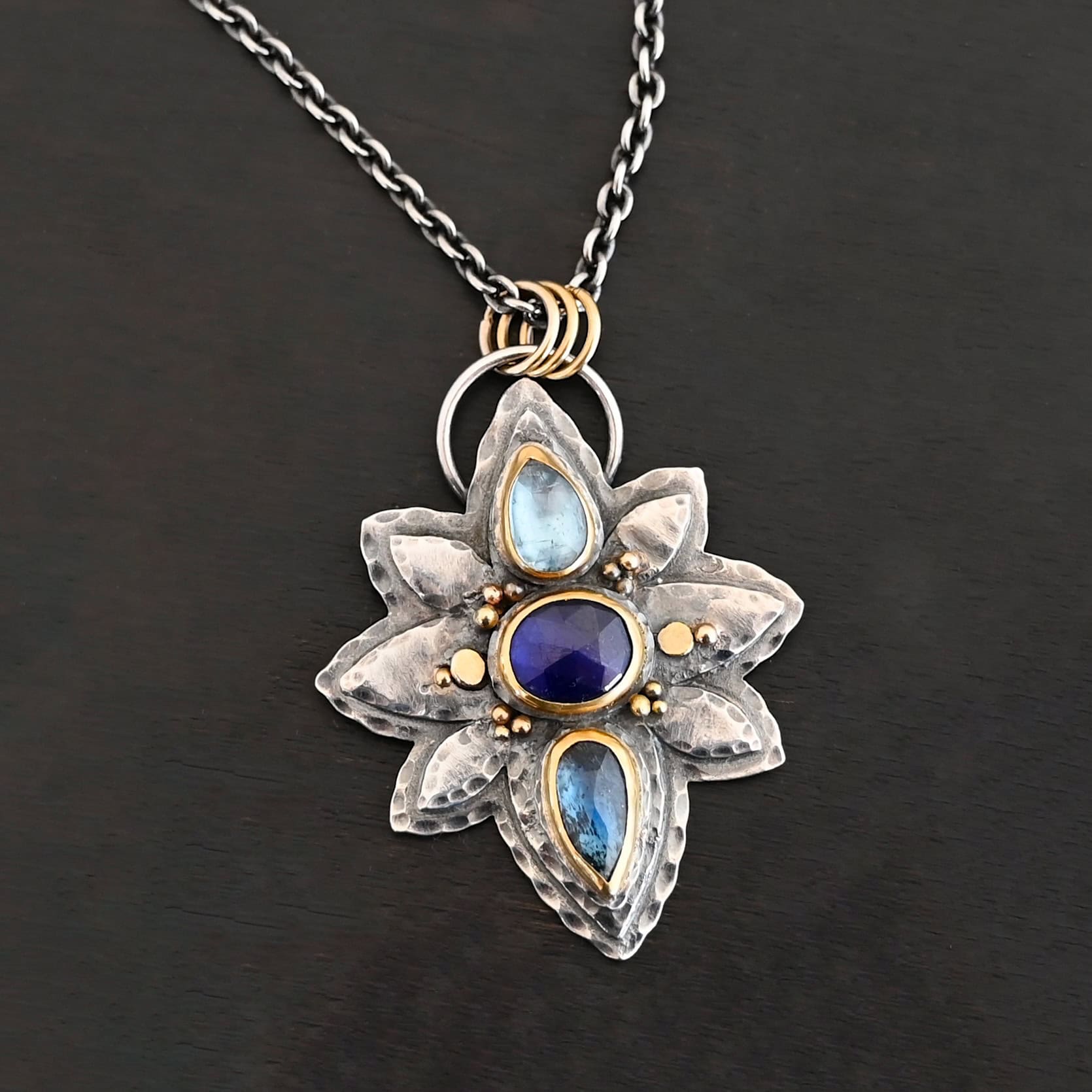 Arendelle Necklace New Photo of the "Arendelle" Necklace. Made out of sterling silver, gold, and tourmaline. There are three tourmalines, each a blue color. The top one is a light blue, and the middle one is a deep royal blue color. The bottom is a dark blue color as well, but not the same as the middle. Pendant is in the shape of a flower with the stones in the center going up and down. There are two layers of sterling silver, each hammered and oxidized. There are three leaf shapes on the top, one in the center and one on each side pointing opposite ways. There is a leaf on each side in the center of the necklace. There are three more leaves on the bottom of the necklace, one in the center and again, one on each side of the center one pointing opposite ways. There are gold dots around the necklace as well. There are fourteen dots and two hammered dots. Seven of the dots are on the left side. They are in groups of three. Three are on the top of the center stone and three are on the bottom. Two dots are in the middle, one that is hammered and one that is not. The hammered one is closer to the stone, and the one that is not is on the side of that one. The other seven are on the right side and follow the same pattern as the left side. There is a loop on the back of the necklace that is connected to three jump rings that are connected to the chain of the necklace. Necklace is laying against a dark background.