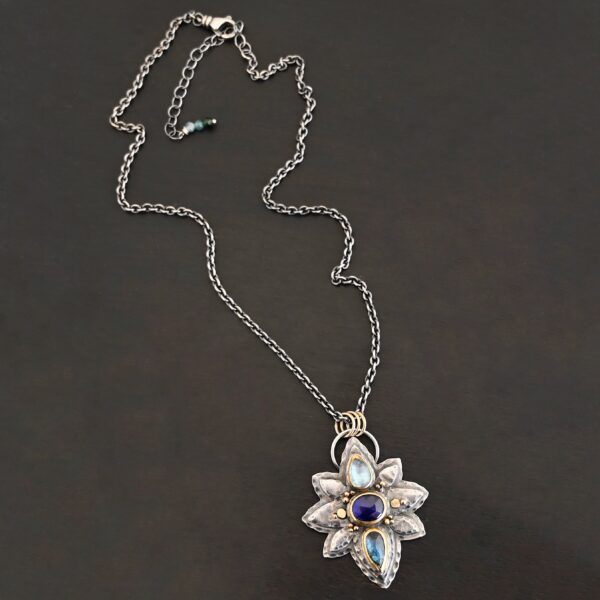 Photo of the "Arendelle" Necklace. Made out of sterling silver, gold, and tourmaline. There are three tourmalines, each a blue color. The top one is a light blue, and the middle one is a deep royal blue color. The bottom is a dark blue color as well, but not the same as the middle. Pendant is in the shape of a flower with the stones in the center going up and down. There are two layers of sterling silver, each hammered and oxidized. There are three leaf shapes on the top, one in the center and one on each side pointing opposite ways. There is a leaf on each side in the center of the necklace. There are three more leaves on the bottom of the necklace, one in the center and again, one on each side of the center one pointing opposite ways. There are gold dots around the necklace as well. There are fourteen dots and two hammered dots. Seven of the dots are on the left side. They are in groups of three. Three are on the top of the center stone and three are on the bottom. Two dots are in the middle, one that is hammered and one that is not. The hammered one is closer to the stone, and the one that is not is on the side of that one. The other seven are on the right side and follow the same pattern as the left side. There is a loop on the back of the necklace that is connected to three jump rings that are connected to the chain of the necklace. Necklace is laying against a dark background.