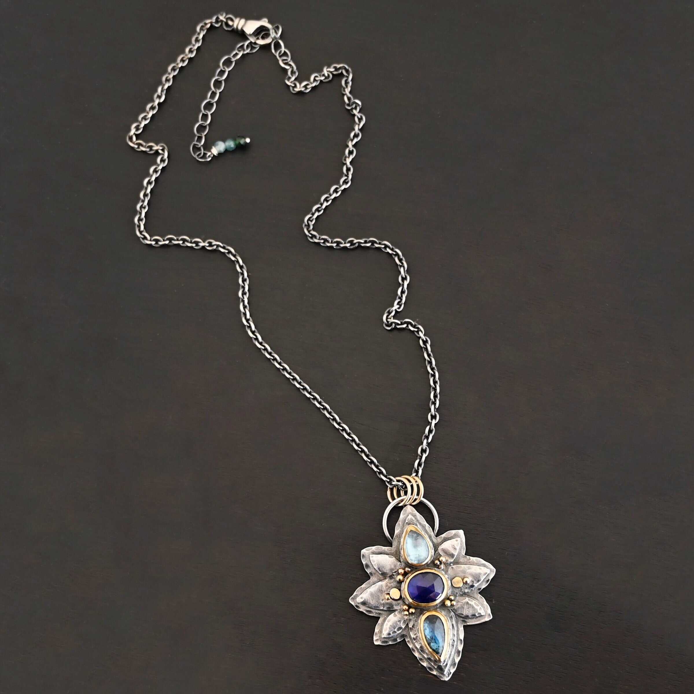 Arendelle Necklace New Photo of the "Arendelle" Necklace. Made out of sterling silver, gold, and tourmaline. There are three tourmalines, each a blue color. The top one is a light blue, and the middle one is a deep royal blue color. The bottom is a dark blue color as well, but not the same as the middle. Pendant is in the shape of a flower with the stones in the center going up and down. There are two layers of sterling silver, each hammered and oxidized. There are three leaf shapes on the top, one in the center and one on each side pointing opposite ways. There is a leaf on each side in the center of the necklace. There are three more leaves on the bottom of the necklace, one in the center and again, one on each side of the center one pointing opposite ways. There are gold dots around the necklace as well. There are fourteen dots and two hammered dots. Seven of the dots are on the left side. They are in groups of three. Three are on the top of the center stone and three are on the bottom. Two dots are in the middle, one that is hammered and one that is not. The hammered one is closer to the stone, and the one that is not is on the side of that one. The other seven are on the right side and follow the same pattern as the left side. There is a loop on the back of the necklace that is connected to three jump rings that are connected to the chain of the necklace. Necklace is laying against a dark background.