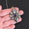 Photo of the "Arendelle" Necklace. Made out of sterling silver, gold, and tourmaline. There are three tourmalines, each a blue color. The top one is a light blue, and the middle one is a deep royal blue color. The bottom is a dark blue color as well, but not the same as the middle. Pendant is in the shape of a flower with the stones in the center going up and down. There are two layers of sterling silver, each hammered and oxidized. There are three leaf shapes on the top, one in the center and one on each side pointing opposite ways. There is a leaf on each side in the center of the necklace. There are three more leaves on the bottom of the necklace, one in the center and again, one on each side of the center one pointing opposite ways. There are gold dots around the necklace as well. There are fourteen dots and two hammered dots. Seven of the dots are on the left side. They are in groups of three. Three are on the top of the center stone and three are on the bottom. Two dots are in the middle, one that is hammered and one that is not. The hammered one is closer to the stone, and the one that is not is on the side of that one. The other seven are on the right side and follow the same pattern as the left side. There is a loop on the back of the necklace that is connected to three jump rings that are connected to the chain of the necklace. Necklace is laying against the fingertips of a hand to demonstrate size of the pendant.