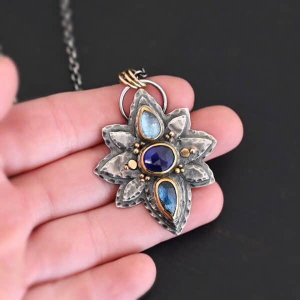 Photo of the "Arendelle" Necklace. Made out of sterling silver, gold, and tourmaline. There are three tourmalines, each a blue color. The top one is a light blue, and the middle one is a deep royal blue color. The bottom is a dark blue color as well, but not the same as the middle. Pendant is in the shape of a flower with the stones in the center going up and down. There are two layers of sterling silver, each hammered and oxidized. There are three leaf shapes on the top, one in the center and one on each side pointing opposite ways. There is a leaf on each side in the center of the necklace. There are three more leaves on the bottom of the necklace, one in the center and again, one on each side of the center one pointing opposite ways. There are gold dots around the necklace as well. There are fourteen dots and two hammered dots. Seven of the dots are on the left side. They are in groups of three. Three are on the top of the center stone and three are on the bottom. Two dots are in the middle, one that is hammered and one that is not. The hammered one is closer to the stone, and the one that is not is on the side of that one. The other seven are on the right side and follow the same pattern as the left side. There is a loop on the back of the necklace that is connected to three jump rings that are connected to the chain of the necklace. Necklace is laying against the fingertips of a hand to demonstrate size of the pendant.