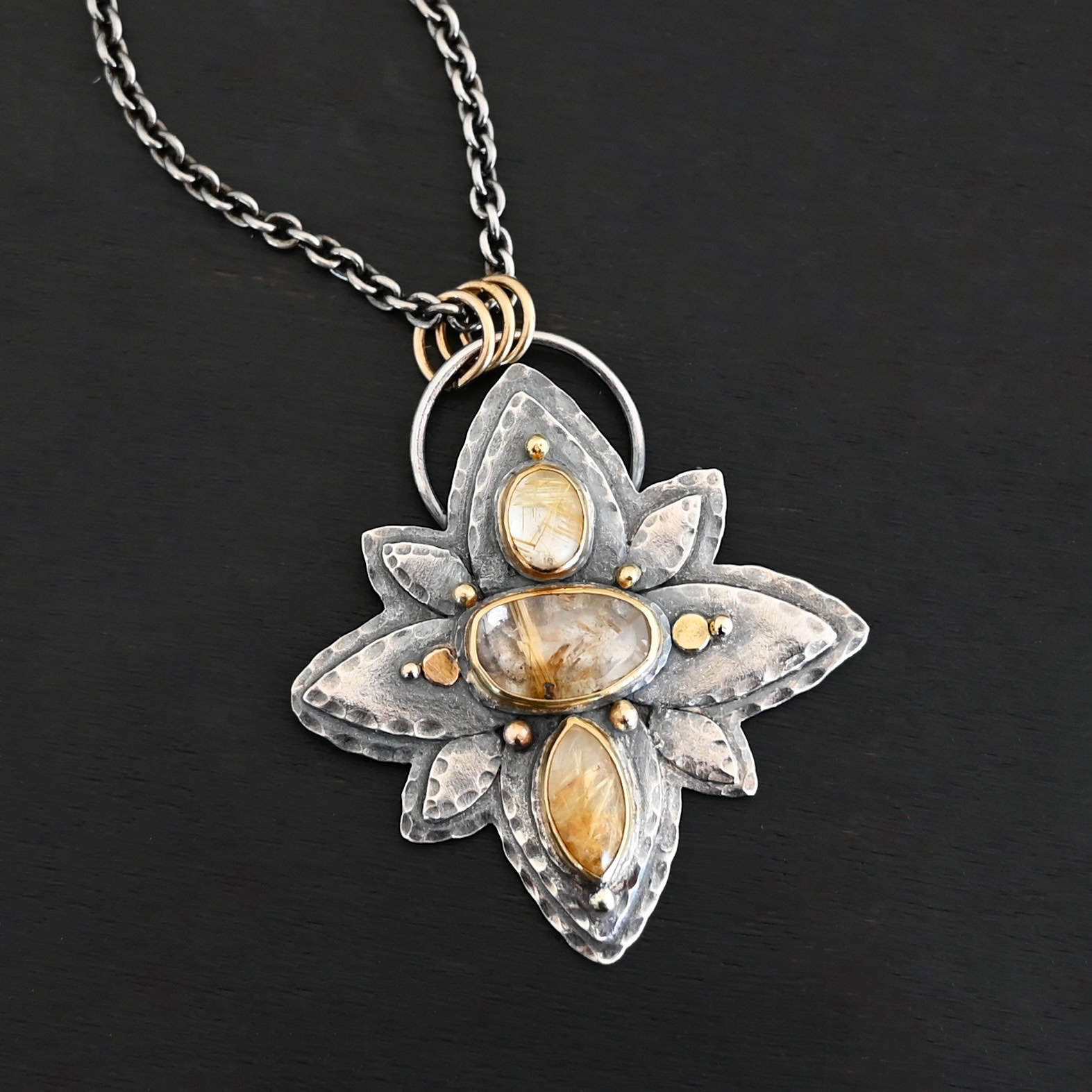 Photo of the "Kingdom Of The Sun" Necklace. Made out of sterling silver, gold, and rutilated quartz. There are three rutilated quartz, each a yellow color with gold specks and lines floating through them. The top one is an oval shape placed up and down. The bottom is a marquise shape. The middle is an oval shape placed sideways. Pendant is in the shape of a flower with the stones in the center going up and down. There are two layers of sterling silver, each hammered and oxidized. There are three leaf shapes on the top, one in the center and one on each side pointing opposite ways. There is a leaf on each side in the center of the necklace. There are three more leaves on the bottom of the necklace, one in the center and again, one on each side of the center one pointing opposite ways. There are gold dots around the necklace as well. There are eight dots and two hammered dots. One of the dots is on the top of the top stone. Two are on each side of the top of the middle stone. There is one on each side of the center stone as well, along with a hammered gold dot on each side of the center stone. There are two more gold dots on the bottom of the center stone on each side. The last gold dot is on the bottom of the marquise shaped stone. There is a loop on the back of the necklace that is connected to three jump rings that are connected to the chain of the necklace. Necklace is laying against a dark background.