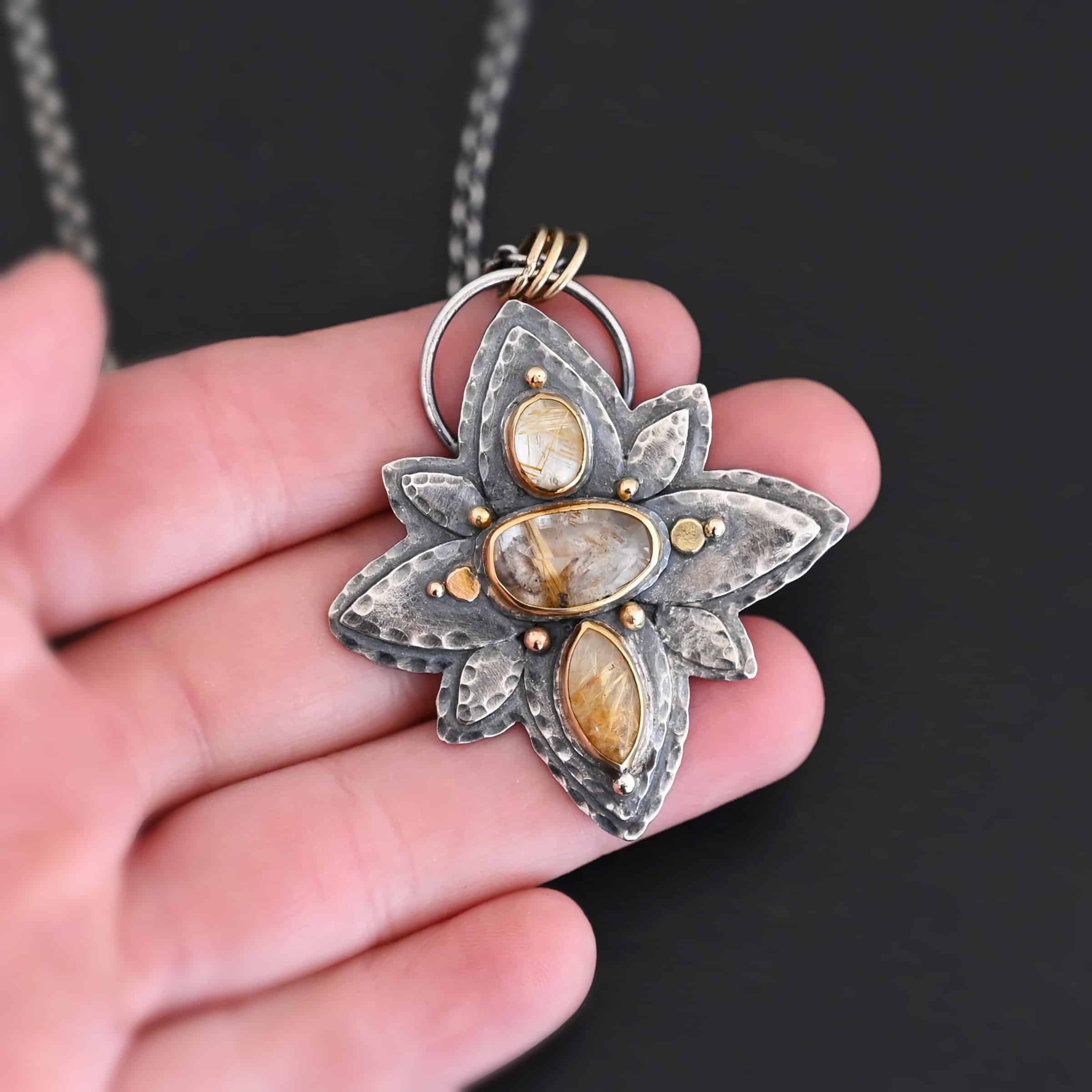Photo of the "Kingdom Of The Sun" Necklace. Made out of sterling silver, gold, and rutilated quartz. There are three rutilated quartz, each a yellow color with gold specks and lines floating through them. The top one is an oval shape placed up and down. The bottom is a marquise shape. The middle is an oval shape placed sideways. Pendant is in the shape of a flower with the stones in the center going up and down. There are two layers of sterling silver, each hammered and oxidized. There are three leaf shapes on the top, one in the center and one on each side pointing opposite ways. There is a leaf on each side in the center of the necklace. There are three more leaves on the bottom of the necklace, one in the center and again, one on each side of the center one pointing opposite ways. There are gold dots around the necklace as well. There are eight dots and two hammered dots. One of the dots is on the top of the top stone. Two are on each side of the top of the middle stone. There is one on each side of the center stone as well, along with a hammered gold dot on each side of the center stone. There are two more gold dots on the bottom of the center stone on each side. The last gold dot is on the bottom of the marquise shaped stone. There is a loop on the back of the necklace that is connected to three jump rings that are connected to the chain of the necklace. Necklace is laying against the fingertips of a hand to demonstrate size of the pendant.