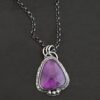 Photo of the "Ought To Do Good" Necklace. Made out of sterling silver and amethyst. The amethyst is a triangle shape but with curved points. The bottom of the amethyst has three silver dots in a straight line that go across the flat bottom. The rest of the silver setting is hammered. There is a loop on the back with three jump rings that are attached to the chain of the necklace. Necklace is laying against a dark background.
