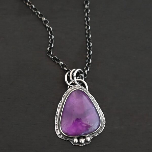 Photo of the "Ought To Do Good" Necklace. Made out of sterling silver and amethyst. The amethyst is a triangle shape but with curved points. The bottom of the amethyst has three silver dots in a straight line that go across the flat bottom. The rest of the silver setting is hammered. There is a loop on the back with three jump rings that are attached to the chain of the necklace. Necklace is laying against a dark background.