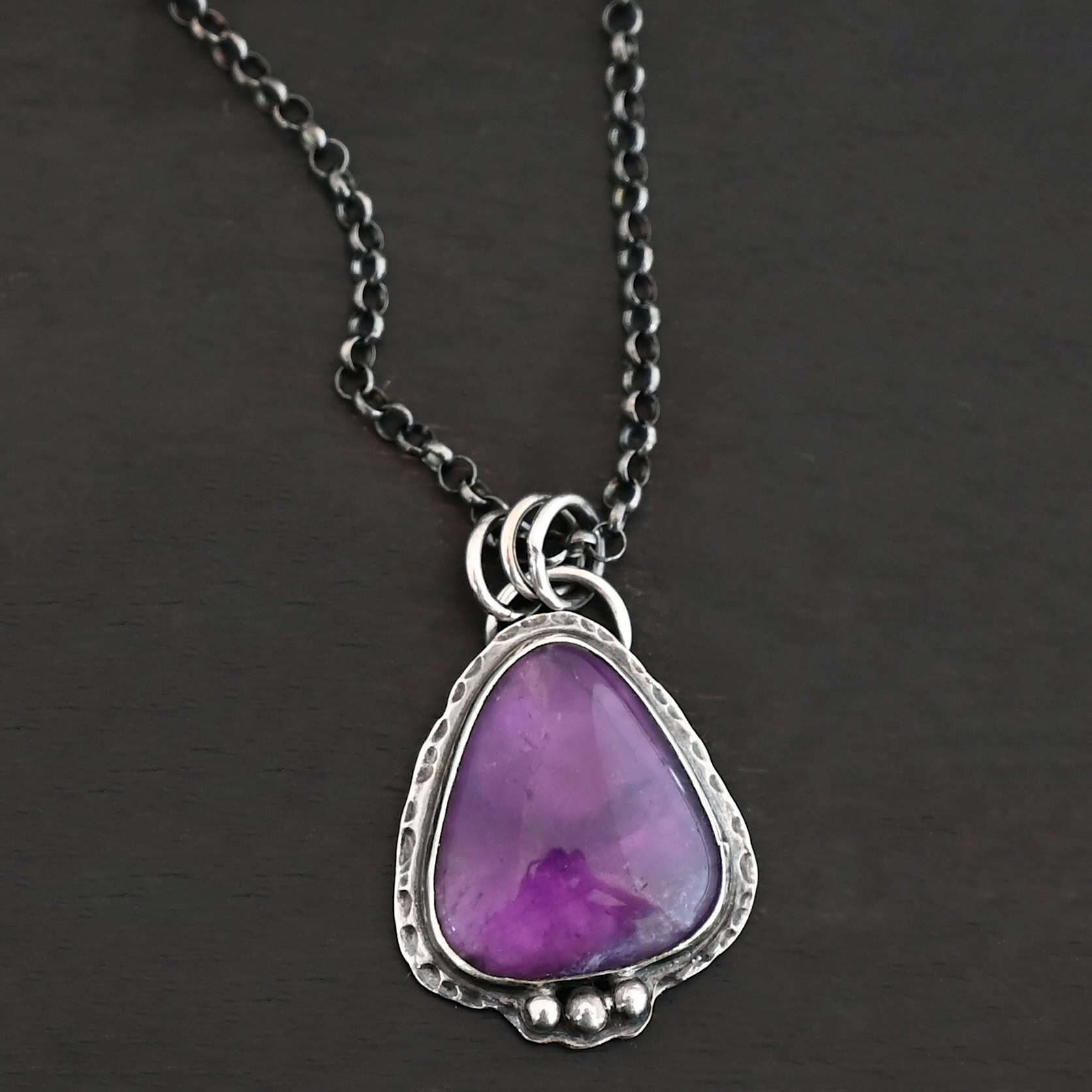 Ought To Do Good Necklace New Photo of the "Ought To Do Good" Necklace. Made out of sterling silver and amethyst. The amethyst is a triangle shape but with curved points. The bottom of the amethyst has three silver dots in a straight line that go across the flat bottom. The rest of the silver setting is hammered. There is a loop on the back with three jump rings that are attached to the chain of the necklace. Necklace is laying against a dark background.