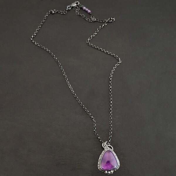 Photo of the "Ought To Do Good" Necklace. Made out of sterling silver and amethyst. The amethyst is a triangle shape but with curved points. The bottom of the amethyst has three silver dots in a straight line that go across the flat bottom. The rest of the silver setting is hammered. There is a loop on the back with three jump rings that are attached to the chain of the necklace. Necklace is laying against a dark background.