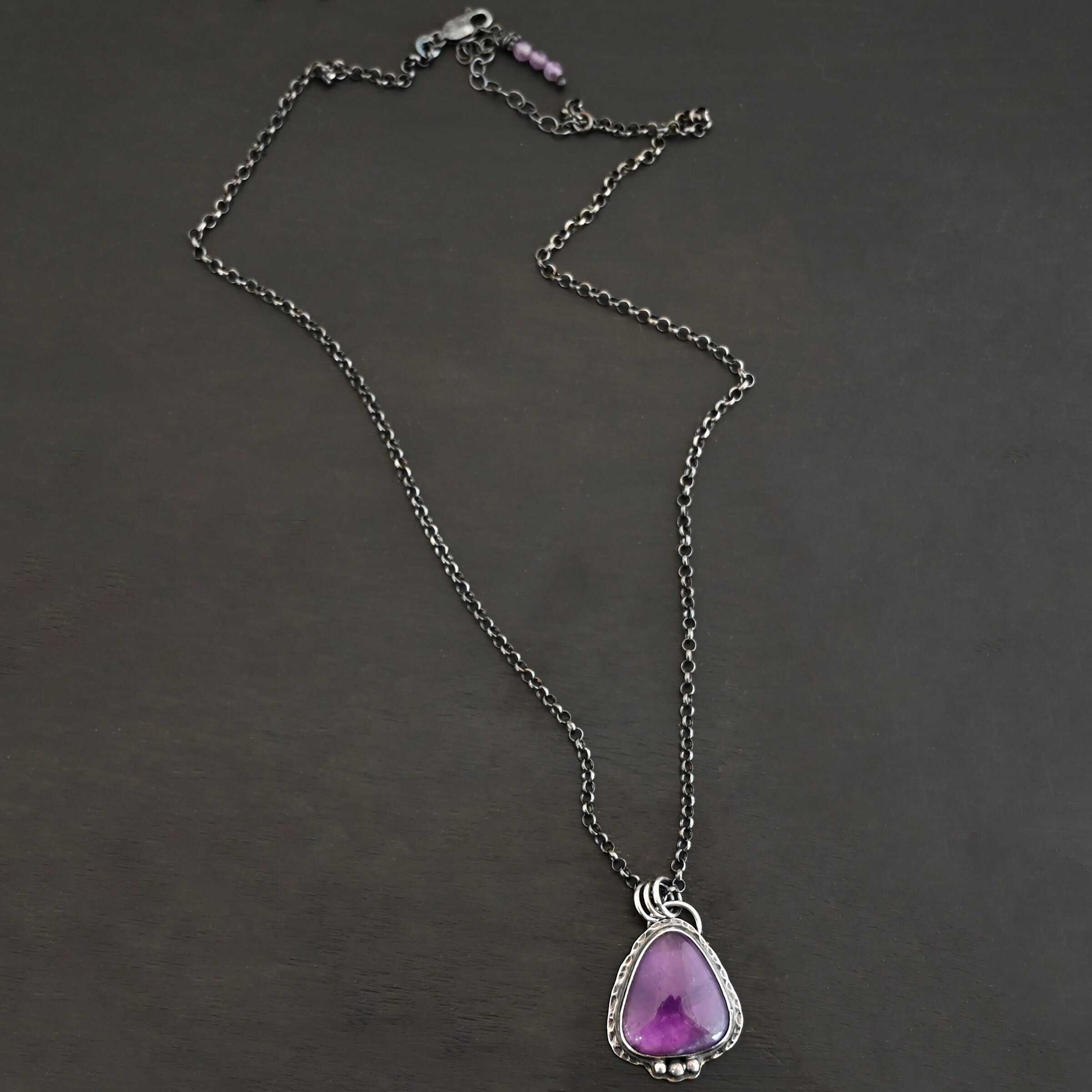 Ought To Do Good Necklace New Photo of the "Ought To Do Good" Necklace. Made out of sterling silver and amethyst. The amethyst is a triangle shape but with curved points. The bottom of the amethyst has three silver dots in a straight line that go across the flat bottom. The rest of the silver setting is hammered. There is a loop on the back with three jump rings that are attached to the chain of the necklace. Necklace is laying against a dark background.