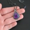 Photo of the "Ought To Do Good" Necklace. Made out of sterling silver and amethyst. The amethyst is a triangle shape but with curved points. The bottom of the amethyst has three silver dots in a straight line that go across the flat bottom. The rest of the silver setting is hammered. There is a loop on the back with three jump rings that are attached to the chain of the necklace. Necklace is laying against the fingertips of a hand to demonstrate size of the pendant.