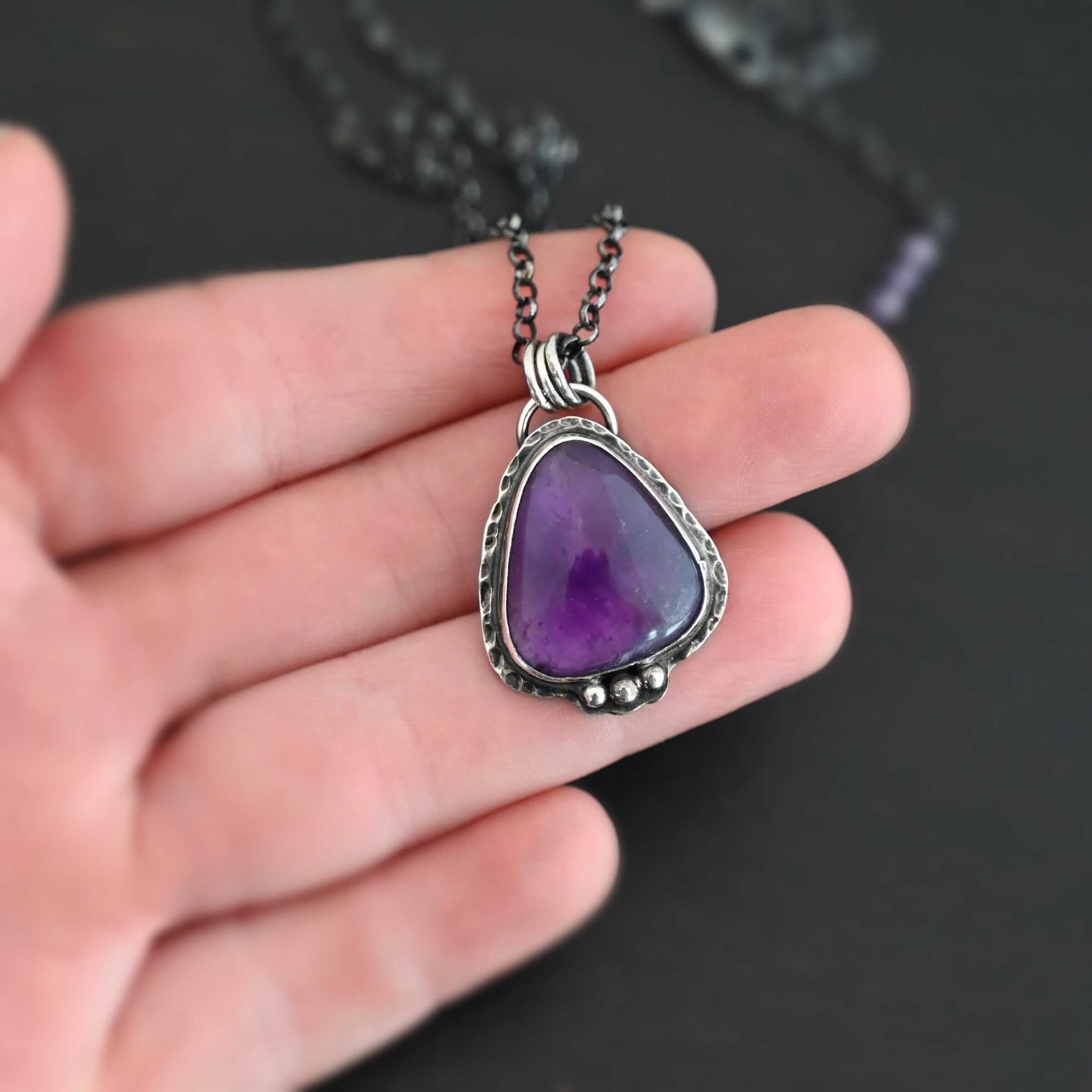Ought To Do Good Necklace New Photo of the "Ought To Do Good" Necklace. Made out of sterling silver and amethyst. The amethyst is a triangle shape but with curved points. The bottom of the amethyst has three silver dots in a straight line that go across the flat bottom. The rest of the silver setting is hammered. There is a loop on the back with three jump rings that are attached to the chain of the necklace. Necklace is laying against the fingertips of a hand to demonstrate size of the pendant.