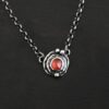 Photo of the "Ciara" Necklace. Made out of sterling silver and tourmaline. The necklace has a small tourmaline in the center, of which is a vibrant red color. The necklace has two layers of sterling silver, and both are hammered. The necklace also has three silver balls scattered around the pendant part of the necklace. There are two loops on the back of the pendant to connect the chain for the necklace to. The necklace is laying against a dark background.