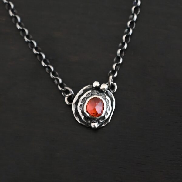 Photo of the "Ciara" Necklace. Made out of sterling silver and tourmaline. The necklace has a small tourmaline in the center, of which is a vibrant red color. The necklace has two layers of sterling silver, and both are hammered. The necklace also has three silver balls scattered around the pendant part of the necklace. There are two loops on the back of the pendant to connect the chain for the necklace to. The necklace is laying against a dark background.