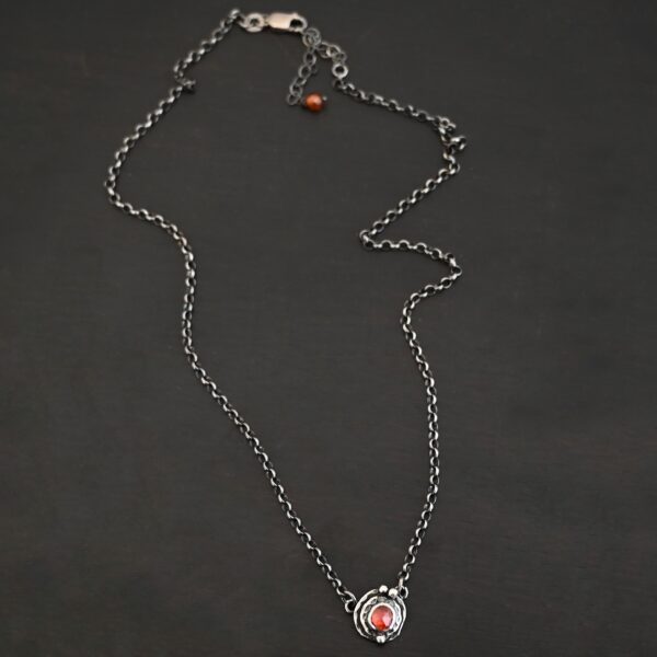 Photo of the "Ciara" Necklace. Made out of sterling silver and tourmaline. The necklace has a small tourmaline in the center, of which is a vibrant red color. The necklace has two layers of sterling silver, and both are hammered. The necklace also has three silver balls scattered around the pendant part of the necklace. There are two loops on the back of the pendant to connect the chain for the necklace to. The necklace is laying against a dark background.
