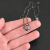 Photo of the "Ciara" Necklace. Made out of sterling silver and tourmaline. The necklace has a small tourmaline in the center, of which is a vibrant red color. The necklace has two layers of sterling silver, and both are hammered. The necklace also has three silver balls scattered around the pendant part of the necklace. There are two loops on the back of the pendant to connect the chain for the necklace to. The necklace is laying in the fingertips of a hand to demonstrate size.