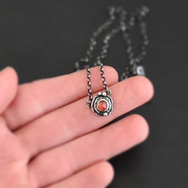 Photo of the "Ciara" Necklace. Made out of sterling silver and tourmaline. The necklace has a small tourmaline in the center, of which is a vibrant red color. The necklace has two layers of sterling silver, and both are hammered. The necklace also has three silver balls scattered around the pendant part of the necklace. There are two loops on the back of the pendant to connect the chain for the necklace to. The necklace is laying in the fingertips of a hand to demonstrate size.