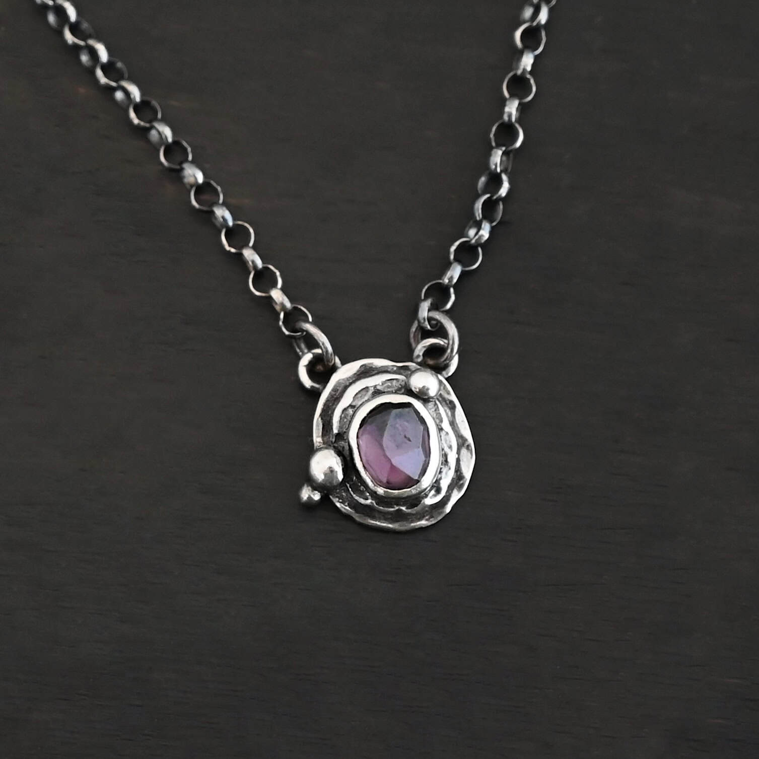 Photo of the "Vera" Necklace. Made out of sterling silver and tourmaline. The necklace has a small tourmaline in the center, of which is a deep purple color. The necklace has two layers of sterling silver, and both are hammered. The necklace also has three silver balls scattered around the pendant part of the necklace. There are two loops on the back of the pendant to connect the chain for the necklace to. The necklace is laying against a dark background.
