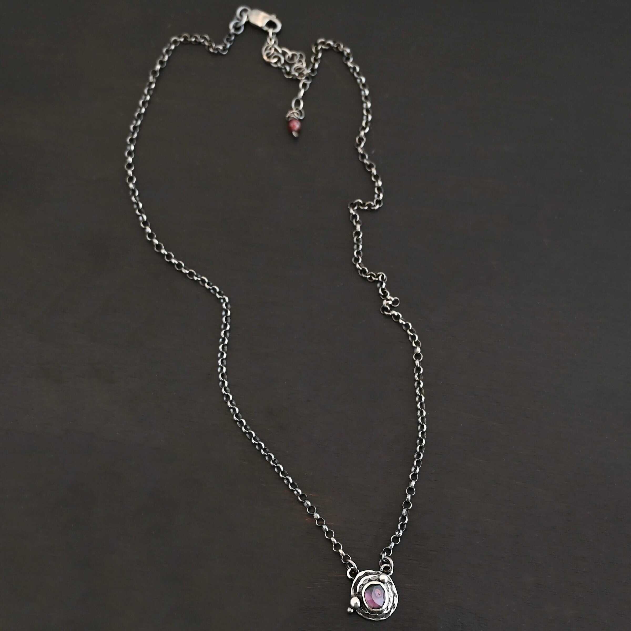 Photo of the "Vera" Necklace. Made out of sterling silver and tourmaline. The necklace has a small tourmaline in the center, of which is a deep purple color. The necklace has two layers of sterling silver, and both are hammered. The necklace also has three silver balls scattered around the pendant part of the necklace. There are two loops on the back of the pendant to connect the chain for the necklace to. The necklace is laying against a dark background.