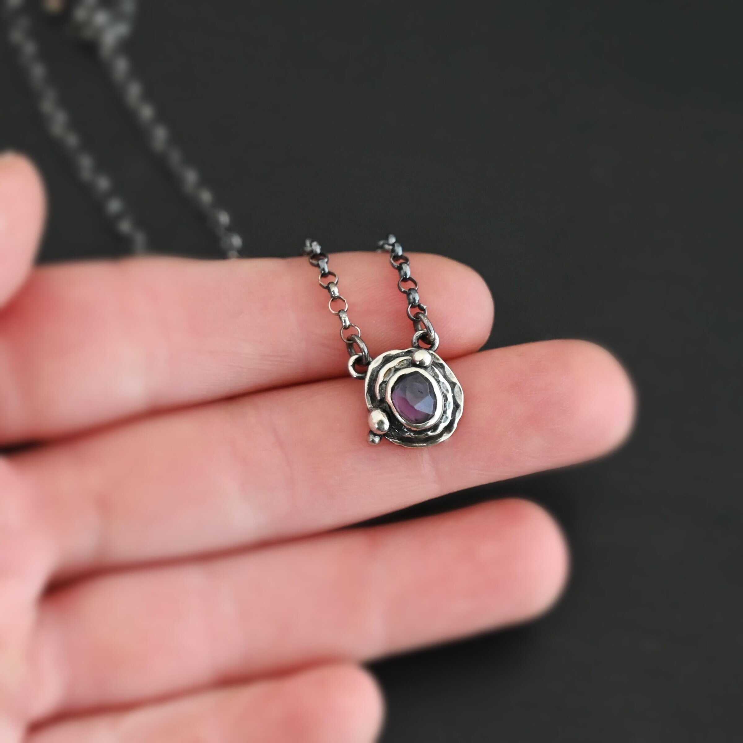 Photo of the "Vera" Necklace. Made out of sterling silver and tourmaline. The necklace has a small tourmaline in the center, of which is a deep purple color. The necklace has two layers of sterling silver, and both are hammered. The necklace also has three silver balls scattered around the pendant part of the necklace. There are two loops on the back of the pendant to connect the chain for the necklace to. The necklace is laying against the fingertips of a hand to demonstrate size of the pendant.