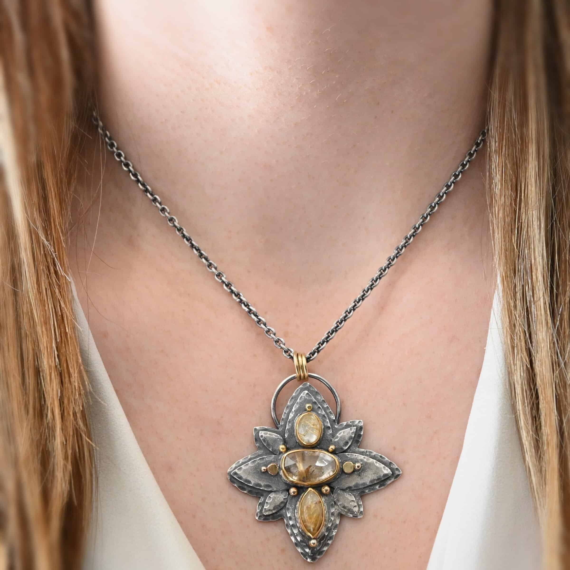 Photo of the "Kingdom Of The Sun" Necklace. Made out of sterling silver, gold, and rutilated quartz. There are three rutilated quartz, each a yellow color with gold specks and lines floating through them. The top one is an oval shape placed up and down. The bottom is a marquise shape. The middle is an oval shape placed sideways. Pendant is in the shape of a flower with the stones in the center going up and down. There are two layers of sterling silver, each hammered and oxidized. There are three leaf shapes on the top, one in the center and one on each side pointing opposite ways. There is a leaf on each side in the center of the necklace. There are three more leaves on the bottom of the necklace, one in the center and again, one on each side of the center one pointing opposite ways. There are gold dots around the necklace as well. There are eight dots and two hammered dots. One of the dots is on the top of the top stone. Two are on each side of the top of the middle stone. There is one on each side of the center stone as well, along with a hammered gold dot on each side of the center stone. There are two more gold dots on the bottom of the center stone on each side. The last gold dot is on the bottom of the marquise shaped stone. There is a loop on the back of the necklace that is connected to three jump rings that are connected to the chain of the necklace. Necklace is worn by a model at 17 inches long to demonstrate size of the pendant.
