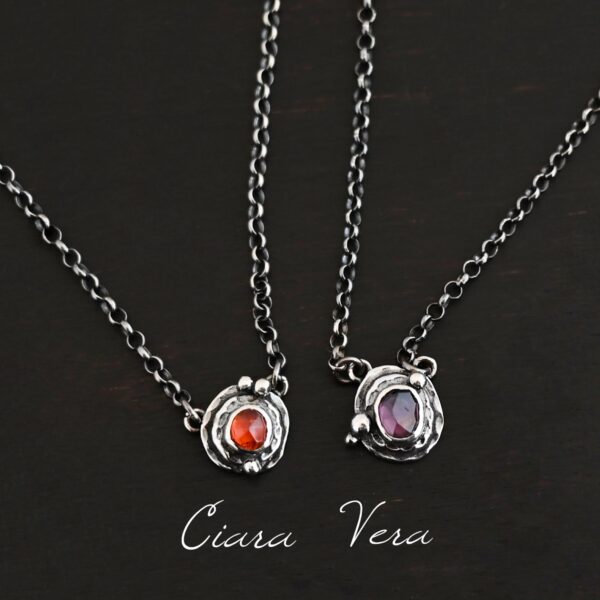 Photo of the "Small But Mighty" Collection. Made out of sterling silver and tourmalines. Each necklace has a small tourmaline in the center, one has a red color, and the other which is a purple color. Each necklace has two layers of sterling silver, and both are hammered. Each necklace also has three silver balls scattered around the pendant part of the necklace. There are two loops on the back of each pendant to connect the chain for the necklace to. From left to right, the necklaces are named Ciara and Vera. Necklaces are laying against a dark background.