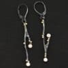 Photo of the "Diamonds In The Trees" Earrings. Made out of sterling silver, gold, and lab created diamonds. Each earring is a sterling silver casted twig off of a branch. There is gold ball detail on each of the twigs. Each earring has leverback ear wires. Each earring has two diamonds. One diamond is on the tip of a small branch near the center. The other diamond is on the end of the twig. Earrings are laying against a dark background.