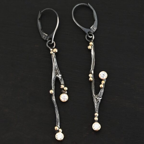 Photo of the "Diamonds In The Trees" Earrings. Made out of sterling silver, gold, and lab created diamonds. Each earring is a sterling silver casted twig off of a branch. There is gold ball detail on each of the twigs. Each earring has leverback ear wires. Each earring has two diamonds. One diamond is on the tip of a small branch near the center. The other diamond is on the end of the twig. Earrings are laying against a dark background.