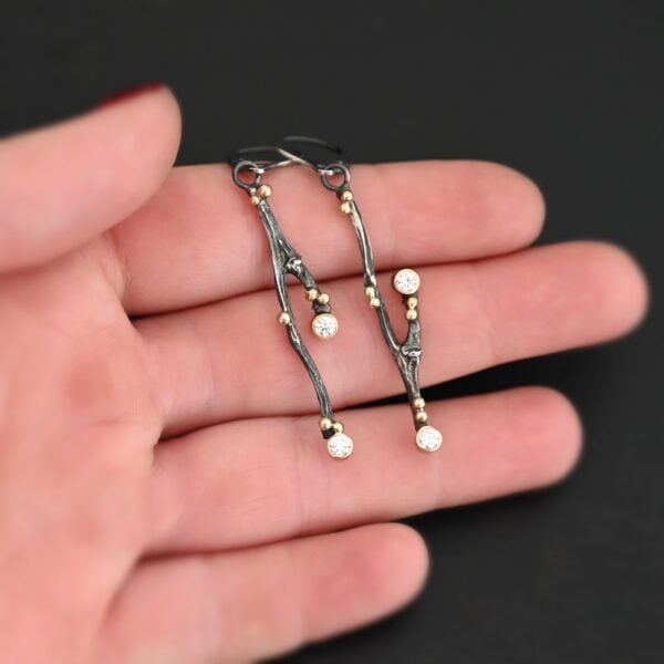 Photo of the "Diamonds In The Trees" Earrings. Made out of sterling silver, gold, and lab created diamonds. Each earring is a sterling silver casted twig off of a branch. There is gold ball detail on each of the twigs. Each earring has leverback ear wires. Each earring has two diamonds. One diamond is on the tip of a small branch near the center. The other diamond is on the end of the twig. Earrings are held in the fingertips of a hand to demonstrate size.