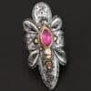 Photo of the "Heaven In A Wildflower" Ring. Made out of sterling silver, gold, ruby, and diamond. There is a marquise shaped ruby in the center of the ring with a small circle diamond beneath it. There is a gold ball on the bottom of the diamond and a hammered silver dot on the top of the marquise. The marquise is placed vertically. There is gold ball detail around the rest of the ring, as well as hammered gold dots. The ring is large and long. The entire ring is shaped like multiple flower petals. There is a large flower petal on top that is rounded, six underneath that flower petal (three on each side), and one on the bottom that is pointed. There are two layers of silver. They are both hammered, but with different patterns. The top leaves on each side and bottom leaves on each side in between the top and bottom leaves have engraved lines in them. Ring is sitting against a dark background.