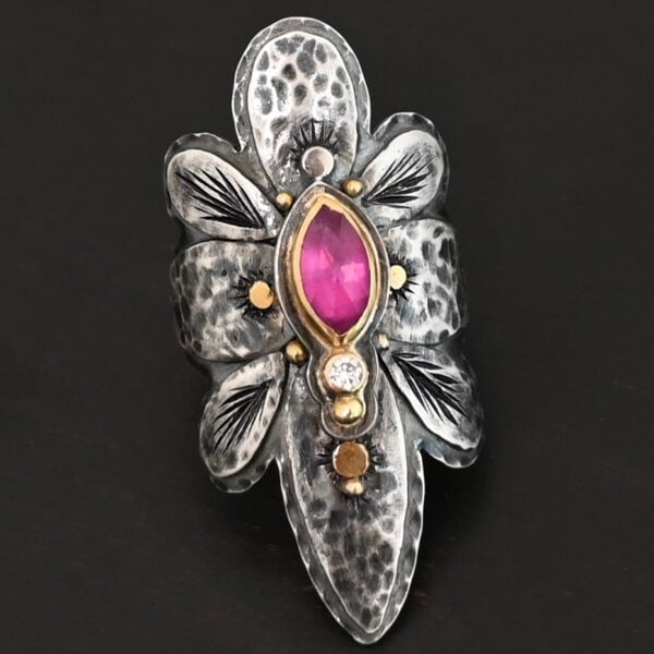 Photo of the "Heaven In A Wildflower" Ring. Made out of sterling silver, gold, ruby, and diamond. There is a marquise shaped ruby in the center of the ring with a small circle diamond beneath it. There is a gold ball on the bottom of the diamond and a hammered silver dot on the top of the marquise. The marquise is placed vertically. There is gold ball detail around the rest of the ring, as well as hammered gold dots. The ring is large and long. The entire ring is shaped like multiple flower petals. There is a large flower petal on top that is rounded, six underneath that flower petal (three on each side), and one on the bottom that is pointed. There are two layers of silver. They are both hammered, but with different patterns. The top leaves on each side and bottom leaves on each side in between the top and bottom leaves have engraved lines in them. Ring is sitting against a dark background.