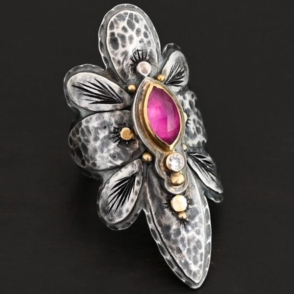 Photo of the "Heaven In A Wildflower" Ring. Made out of sterling silver, gold, ruby, and diamond. There is a marquise shaped ruby in the center of the ring with a small circle diamond beneath it. There is a gold ball on the bottom of the diamond and a hammered silver dot on the top of the marquise. The marquise is placed vertically. There is gold ball detail around the rest of the ring, as well as hammered gold dots. The ring is large and long. The entire ring is shaped like multiple flower petals. There is a large flower petal on top that is rounded, six underneath that flower petal (three on each side), and one on the bottom that is pointed. There are two layers of silver. They are both hammered, but with different patterns. The top leaves on each side and bottom leaves on each side in between the top and bottom leaves have engraved lines in them. Ring is sitting against a dark background.