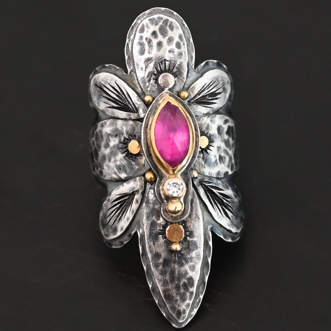 Heaven In A Wildflower Ring Photo of the "Heaven In A Wildflower" Ring. Made out of sterling silver, gold, ruby, and diamond. There is a marquise shaped ruby in the center of the ring with a small circle diamond beneath it. There is a gold ball on the bottom of the diamond and a hammered silver dot on the top of the marquise. The marquise is placed vertically. There is gold ball detail around the rest of the ring, as well as hammered gold dots. The ring is large and long. The entire ring is shaped like multiple flower petals. There is a large flower petal on top that is rounded, six underneath that flower petal (three on each side), and one on the bottom that is pointed. There are two layers of silver. They are both hammered, but with different patterns. The top leaves on each side and bottom leaves on each side in between the top and bottom leaves have engraved lines in them. Ring is sitting against a dark background.