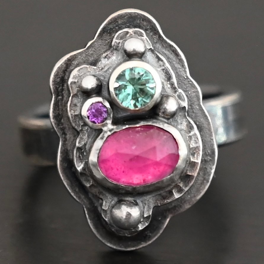 Photo of the "Strength And Softness" Ring. Made out of sterling silver, ruby, amethyst, and green tourmaline. The bottom stone is an oval ruby set vertically. Above this on the left side is a small circle amethyst. To the right side of this, on top of the ruby is a larger green tourmaline that is circular. There is silver ball detail scattered around the stones. There are two layers of sterling silver. The top layer is where all the stones are placed, and this layer is hammered. The bottom layer is larger so it can be seen, but it is not hammered. Ring is sitting against a dark background.