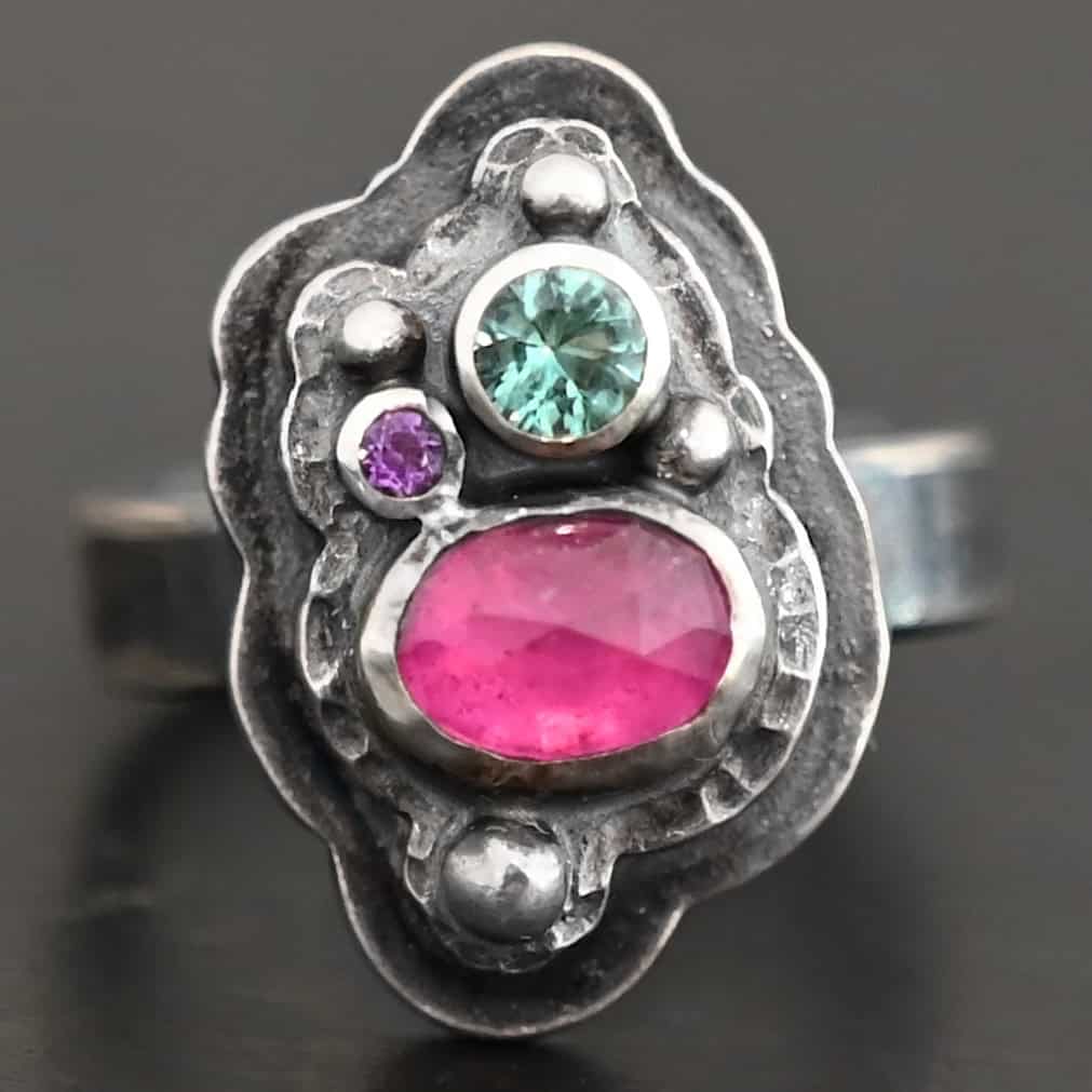 Photo of the "Strength And Softness" Ring. Made out of sterling silver, ruby, amethyst, and green tourmaline. The bottom stone is an oval ruby set vertically. Above this on the left side is a small circle amethyst. To the right side of this, on top of the ruby is a larger green tourmaline that is circular. There is silver ball detail scattered around the stones. There are two layers of sterling silver. The top layer is where all the stones are placed, and this layer is hammered. The bottom layer is larger so it can be seen, but it is not hammered. Ring is sitting against a dark background.