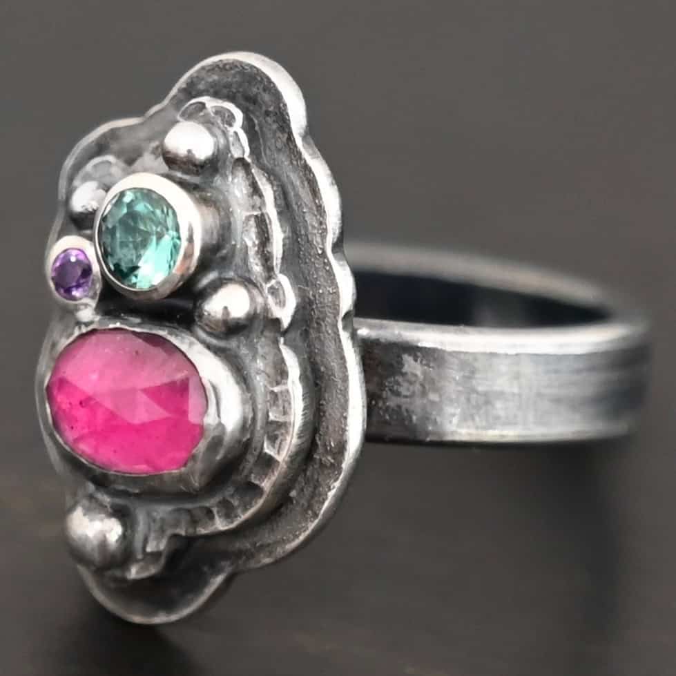 Photo of the "Strength And Softness" Ring. Made out of sterling silver, ruby, amethyst, and green tourmaline. The bottom stone is an oval ruby set vertically. Above this on the left side is a small circle amethyst. To the right side of this, on top of the ruby is a larger green tourmaline that is circular. There is silver ball detail scattered around the stones. There are two layers of sterling silver. The top layer is where all the stones are placed, and this layer is hammered. The bottom layer is larger so it can be seen, but it is not hammered. Ring is sitting against a dark background.