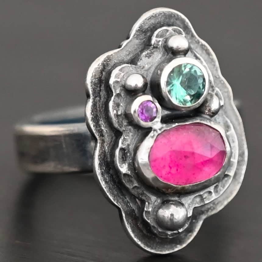 Photo of the "Strength And Softness" Ring. Made out of sterling silver, ruby, amethyst, and green tourmaline. The bottom stone is an oval ruby set vertically. Above this on the left side is a small circle amethyst. To the right side of this, on top of the ruby is a larger green tourmaline that is circular. There is silver ball detail scattered around the stones. There are two layers of sterling silver. The top layer is where all the stones are placed, and this layer is hammered. The bottom layer is larger so it can be seen, but it is not hammered. Ring is sitting against a dark background.