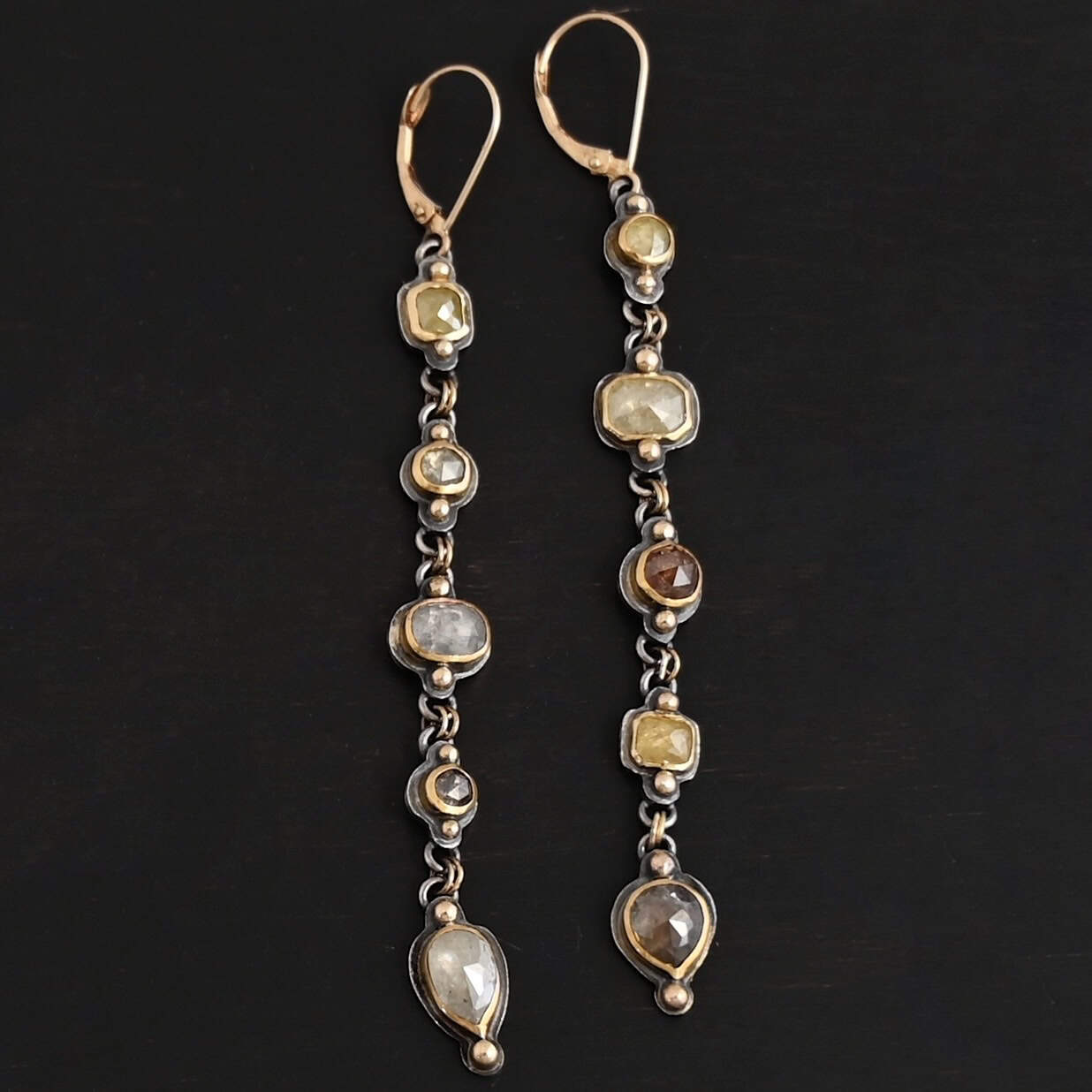 Photo of the "First Light" Earrings. Made out of sterling silver, gold, and diamonds. The top of the earrings are gold leverback ear wires that are connected to small silver loops on the back of the top of the earrings. Each earring has five diamonds that all hang down. Each diamond is set in gold, and the back of each diamond has a loop on the top and bottom to connect it to the previous diamond and the next diamond. Each diamond has two gold balls on the front, one on the top and one on the bottom. Each diamond is connected by two gold jump rings that go through the loops on the back. Earrings are lying against a dark background.