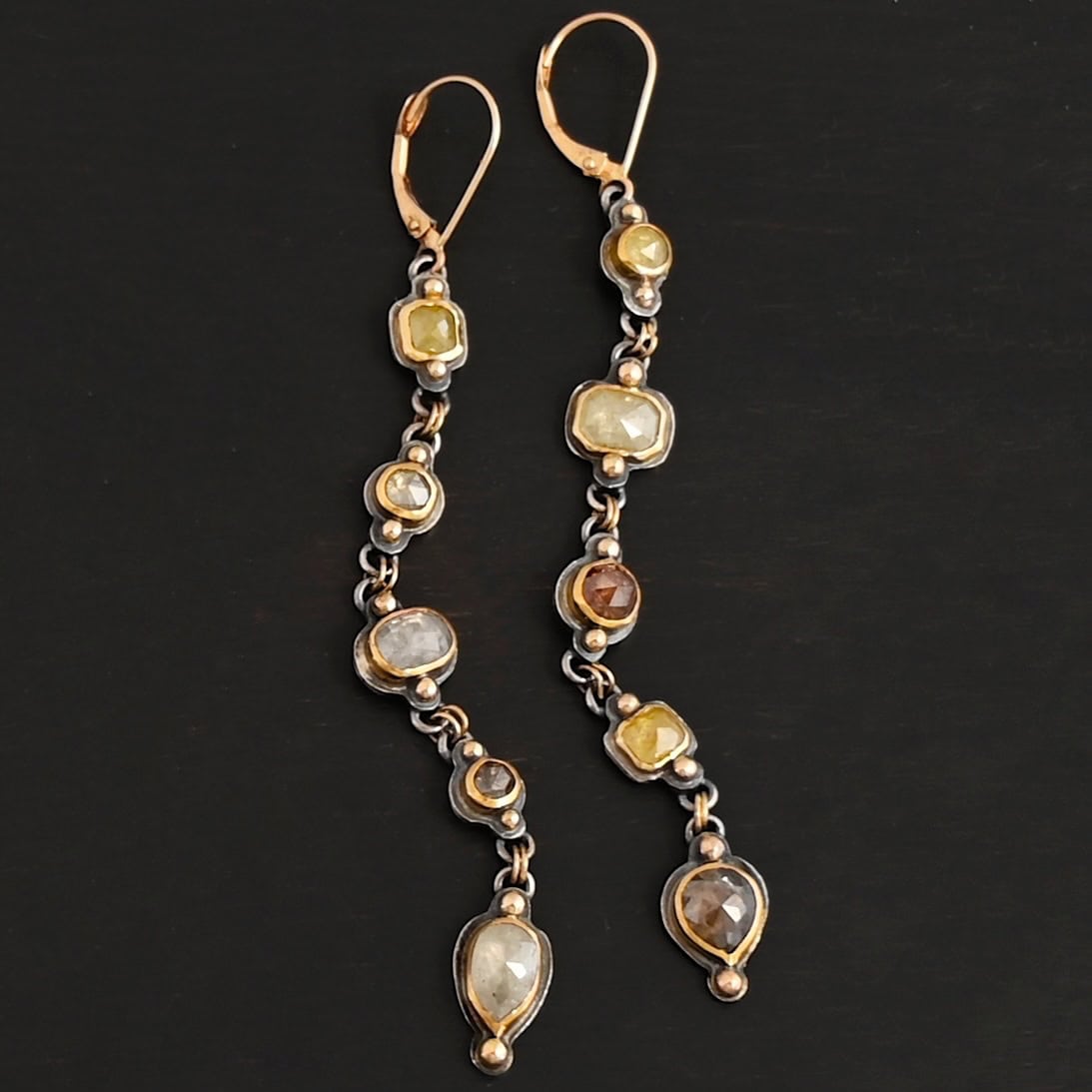 Photo of the "First Light" Earrings. Made out of sterling silver, gold, and diamonds. The top of the earrings are gold leverback ear wires that are connected to small silver loops on the back of the top of the earrings. Each earring has five diamonds that all hang down. Each diamond is set in gold, and the back of each diamond has a loop on the top and bottom to connect it to the previous diamond and the next diamond. Each diamond has two gold balls on the front, one on the top and one on the bottom. Each diamond is connected by two gold jump rings that go through the loops on the back. Earrings are lying against a dark background.