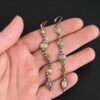 Photo of the "First Light" Earrings. Made out of sterling silver, gold, and diamonds. The top of the earrings are gold leverback ear wires that are connected to small silver loops on the back of the top of the earrings. Each earring has five diamonds that all hang down. Each diamond is set in gold, and the back of each diamond has a loop on the top and bottom to connect it to the previous diamond and the next diamond. Each diamond has two gold balls on the front, one on the top and one on the bottom. Each diamond is connected by two gold jump rings that go through the loops on the back. Earrings are lying against the fingertips of a hand.