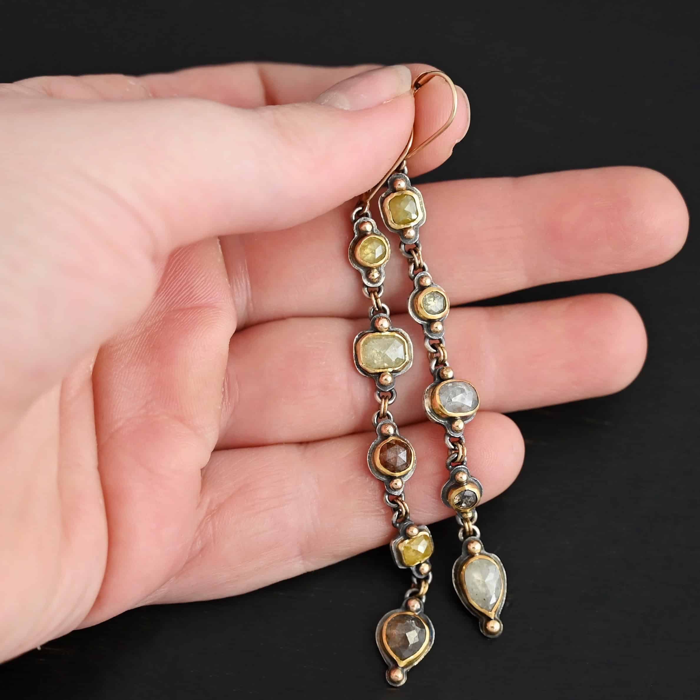 Photo of the "First Light" Earrings. Made out of sterling silver, gold, and diamonds. The top of the earrings are gold leverback ear wires that are connected to small silver loops on the back of the top of the earrings. Each earring has five diamonds that all hang down. Each diamond is set in gold, and the back of each diamond has a loop on the top and bottom to connect it to the previous diamond and the next diamond. Each diamond has two gold balls on the front, one on the top and one on the bottom. Each diamond is connected by two gold jump rings that go through the loops on the back. Earrings are lying against the fingertips of a hand.