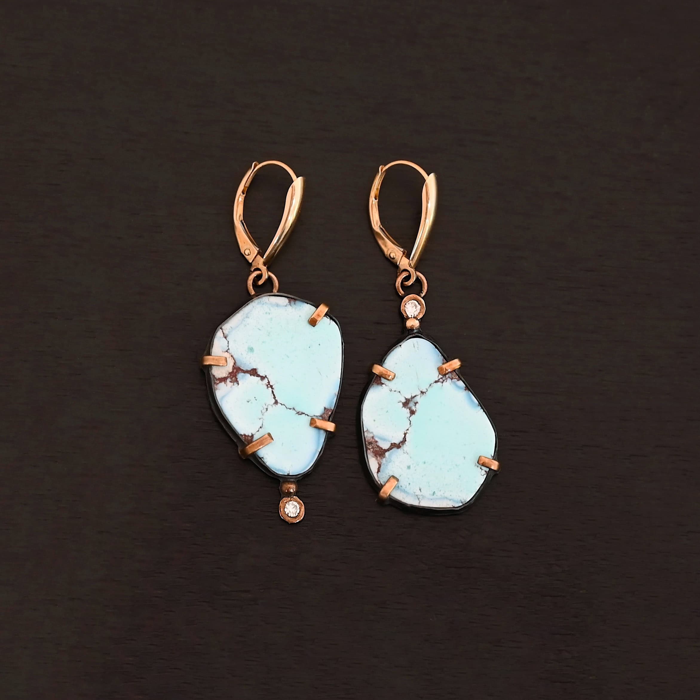 Path Of A Glacier Earrings New Photo of the "Path Of A Glacier" Earrings. Made out of sterling silver, gold, turquoise, and lab created diamonds. Both earrings have a large oval shaped turquoise in the center with four gold prongs, two per side, holding the stones in. There is a gold ball on the top of the turquoise on the left earring and a gold ball on the bottom turquoise of the right earring. The left earring has a diamond on the top of the gold ball and the right earring has the diamond on the bottom of the gold ball. At the top of each earring, on the back of it is a gold loop. Attached to these loops is a gold leverback ear wire. Earrings are laying against a dark background.
