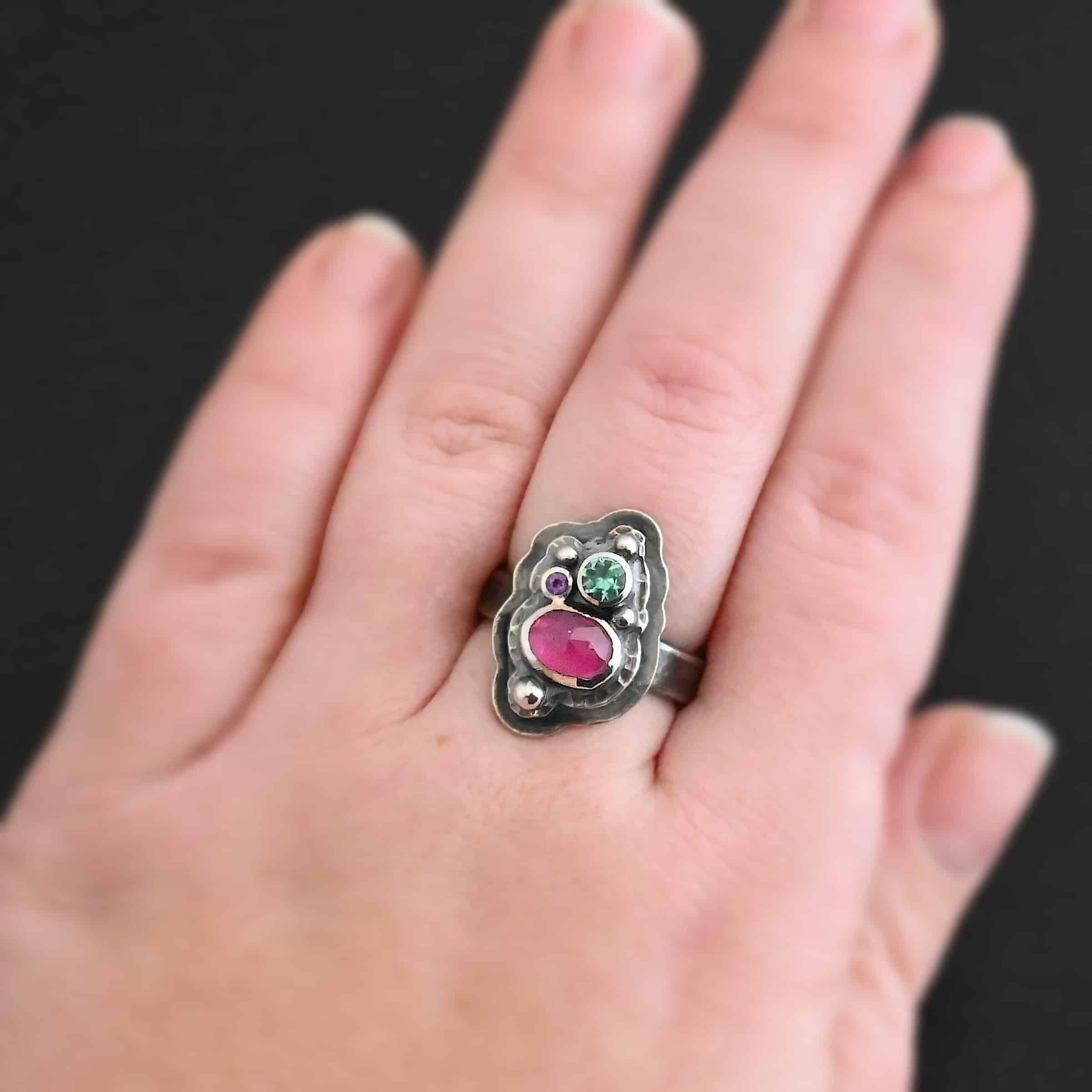 Photo of the "Strength And Softness" Ring. Made out of sterling silver, ruby, amethyst, and green tourmaline. The bottom stone is an oval ruby set vertically. Above this on the left side is a small circle amethyst. To the right side of this, on top of the ruby is a larger green tourmaline that is circular. There is silver ball detail scattered around the stones. There are two layers of sterling silver. The top layer is where all the stones are placed, and this layer is hammered. The bottom layer is larger so it can be seen, but it is not hammered. Ring is worn by a model up close to demonstrate size on their engagement ring finger.