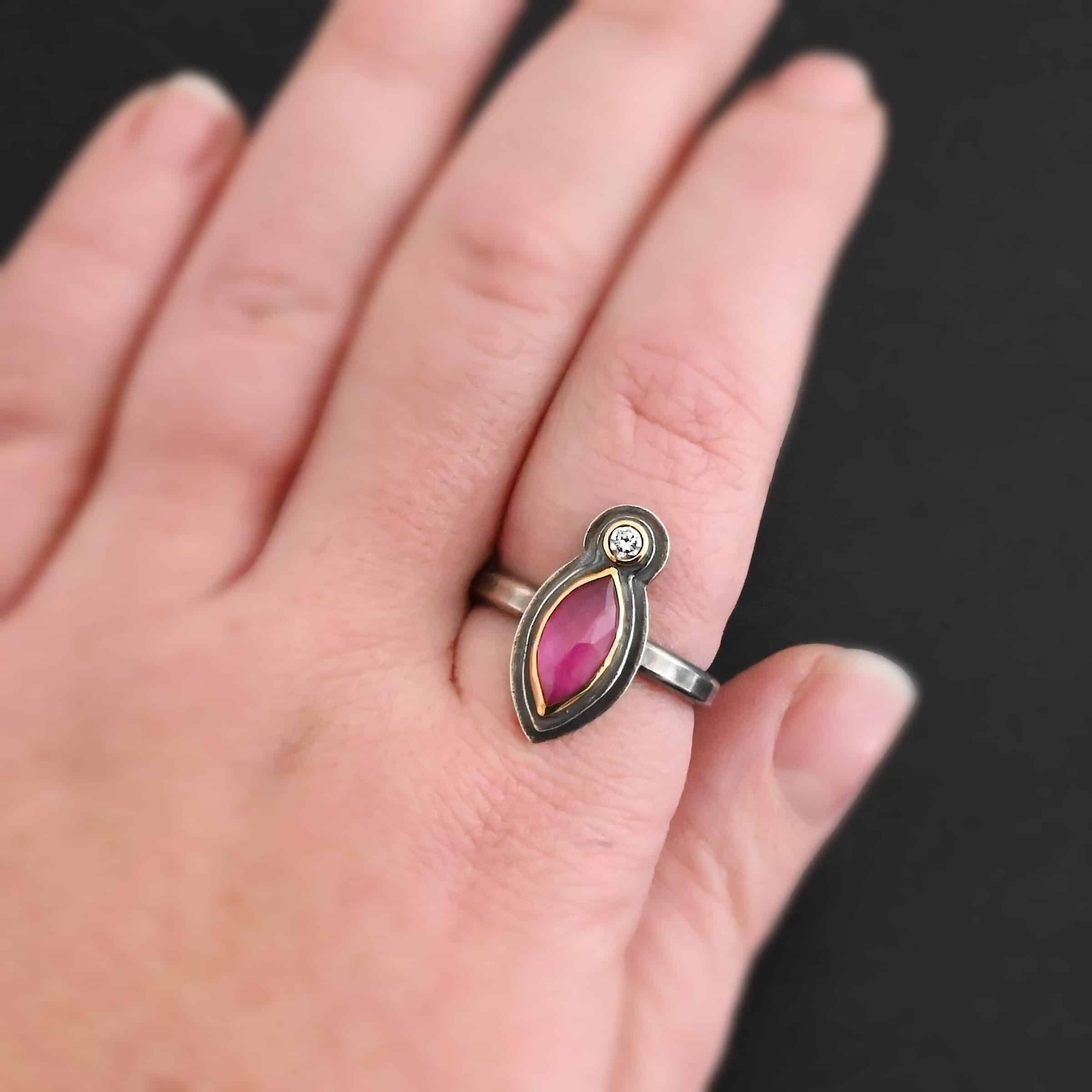 Soft Petals Ring Photo of the "Soft Petals" Ring. Made out of sterling silver, gold, ruby, and diamond. The top of the ring has a small circle diamond. Beneath this is a pink, marquise shaped ruby placed vertically, so that the points are pointing up and down. The stone is set in gold, and there are two layers of sterling silver beneath it. The bottom layer is bigger than the top layer of silver so that it is noticeable. The ring band is a smooth band that is thin. Ring is worn up close by a model to demonstrate size.