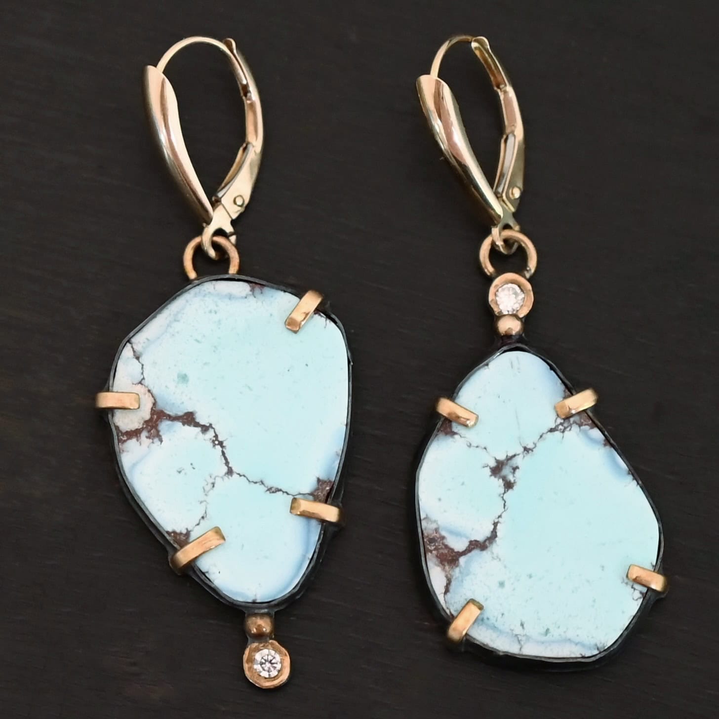 Path Of A Glacier Earrings New Photo of the "Path Of A Glacier" Earrings. Made out of sterling silver, gold, turquoise, and lab created diamonds. Both earrings have a large oval shaped turquoise in the center with four gold prongs, two per side, holding the stones in. There is a gold ball on the top of the turquoise on the left earring and a gold ball on the bottom turquoise of the right earring. The left earring has a diamond on the top of the gold ball and the right earring has the diamond on the bottom of the gold ball. At the top of each earring, on the back of it is a gold loop. Attached to these loops is a gold leverback ear wire. Earrings are laying against a dark background.
