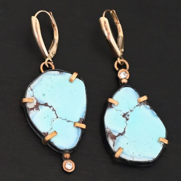 Photo of the "Path Of A Glacier" Earrings. Made out of sterling silver, gold, turquoise, and lab created diamonds. Both earrings have a large oval shaped turquoise in the center with four gold prongs, two per side, holding the stones in. There is a gold ball on the top of the turquoise on the left earring and a gold ball on the bottom turquoise of the right earring. The left earring has a diamond on the top of the gold ball and the right earring has the diamond on the bottom of the gold ball. At the top of each earring, on the back of it is a gold loop. Attached to these loops is a gold leverback ear wire. Earrings are laying against a dark background.
