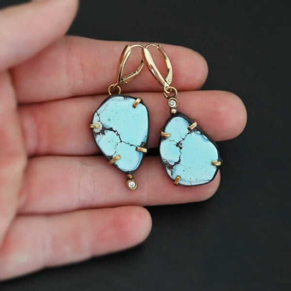 Photo of the "Path Of A Glacier" Earrings. Made out of sterling silver, gold, turquoise, and lab created diamonds. Both earrings have a large oval shaped turquoise in the center with four gold prongs, two per side, holding the stones in. There is a gold ball on the top of the turquoise on the left earring and a gold ball on the bottom turquoise of the right earring. The left earring has a diamond on the top of the gold ball and the right earring has the diamond on the bottom of the gold ball. At the top of each earring, on the back of it is a gold loop. Attached to these loops is a gold leverback ear wire. Earrings are laying against the fingertips of a hand to demonstrate size.