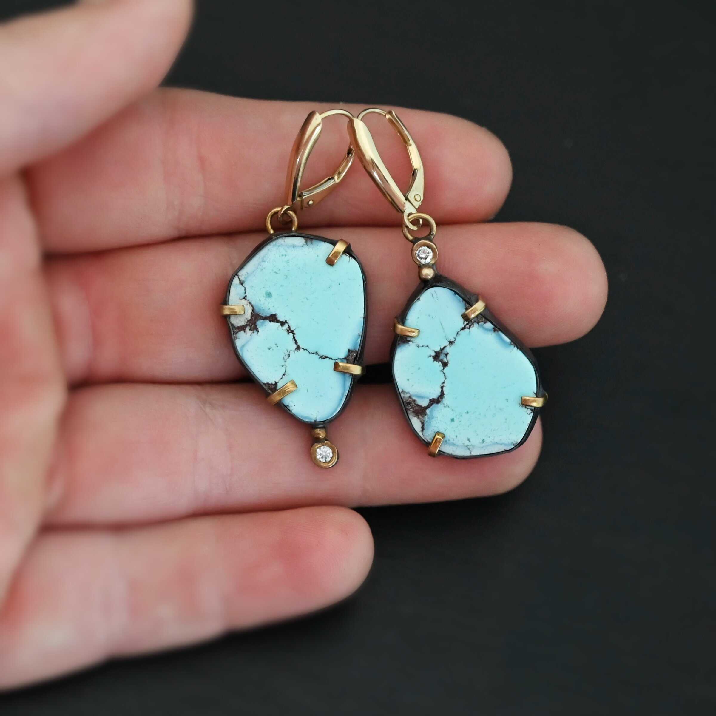 Path Of A Glacier Earrings New Photo of the "Path Of A Glacier" Earrings. Made out of sterling silver, gold, turquoise, and lab created diamonds. Both earrings have a large oval shaped turquoise in the center with four gold prongs, two per side, holding the stones in. There is a gold ball on the top of the turquoise on the left earring and a gold ball on the bottom turquoise of the right earring. The left earring has a diamond on the top of the gold ball and the right earring has the diamond on the bottom of the gold ball. At the top of each earring, on the back of it is a gold loop. Attached to these loops is a gold leverback ear wire. Earrings are laying against the fingertips of a hand to demonstrate size.