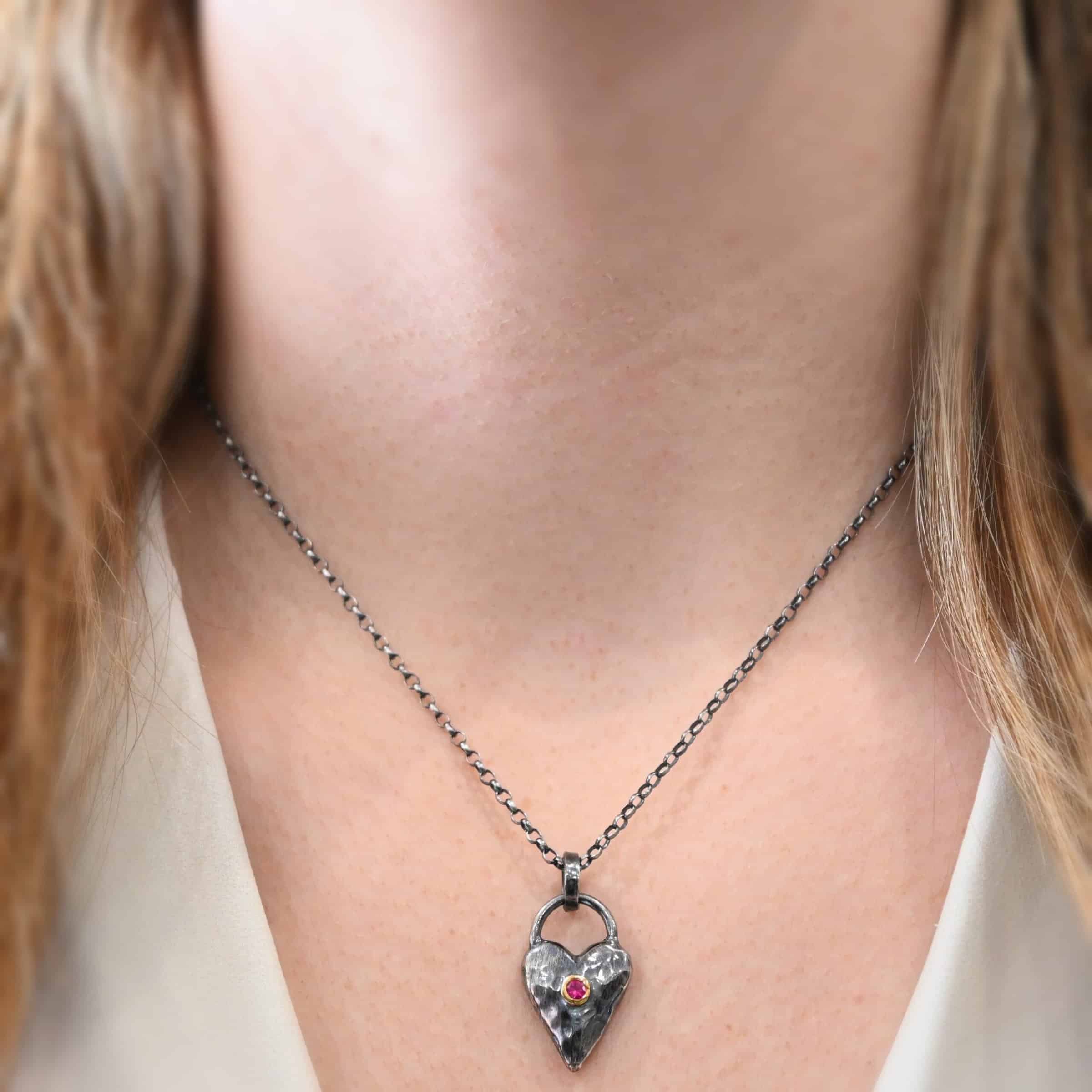 In Your Heart Birthstone Necklace - Ruby Photo of the "In Your Heart" Necklace. Made out of sterling silver and gold with a birthstone. The pendant is a solid sterling silver heart that is hammered and oxidized with a ruby birthstone in the center that is set in gold. There is a silver loop that goes from one side of the heart to the other on the top. Attached to this heart is a large jump ring that is oxidized. Going through this is the silver chain for the necklace. Necklace is worn by a model up close at 17" long to demonstrate how it looks.