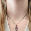 Photo of the "Soft Petals" Necklace. Made out of sterling silver, gold, ruby, and diamond. The bottom of the ring has a small circle diamond. Above this is a pink, marquise shaped ruby placed vertically, so that the points are pointing up and down. The stone is set in gold, and there are two layers of sterling silver beneath it. The top layer of silver goes around the edge of the bottom layer. Both layers are marquise shaped, and the top outside layer is hammered. There is a loop on the back of the necklace in which three gold jump rings go through. Going through the jump rings is the silver chain of the necklace. Necklace is worn by a model at 16" long to demonstrate size.