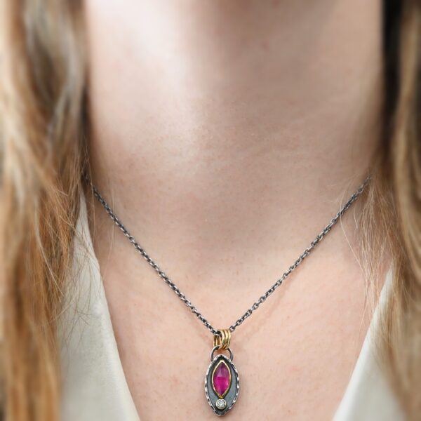 Photo of the "Soft Petals" Necklace. Made out of sterling silver, gold, ruby, and diamond. The bottom of the ring has a small circle diamond. Above this is a pink, marquise shaped ruby placed vertically, so that the points are pointing up and down. The stone is set in gold, and there are two layers of sterling silver beneath it. The top layer of silver goes around the edge of the bottom layer. Both layers are marquise shaped, and the top outside layer is hammered. There is a loop on the back of the necklace in which three gold jump rings go through. Going through the jump rings is the silver chain of the necklace. Necklace is worn by a model at 16" long to demonstrate size.