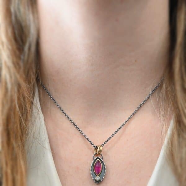 Photo of the "Soft Petals" Necklace. Made out of sterling silver, gold, ruby, and diamond. The bottom of the ring has a small circle diamond. Above this is a pink, marquise shaped ruby placed vertically, so that the points are pointing up and down. The stone is set in gold, and there are two layers of sterling silver beneath it. The top layer of silver goes around the edge of the bottom layer. Both layers are marquise shaped, and the top outside layer is hammered. There is a loop on the back of the necklace in which three gold jump rings go through. Going through the jump rings is the silver chain of the necklace. Necklace is worn by a model at 16" long to demonstrate size.
